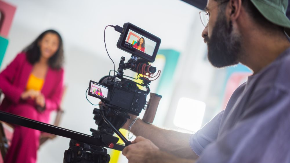 Film Set With A Young Female Host In A Pink Suit Engaging In A Lively Discussion, Captured By A Male Cinematographer Focusing Intently On His Camera Equipment, Surrounded By Colorful Backdrops.