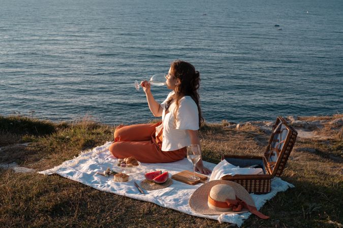 Woman with glass of drink on picnic mat looking at sea and mountains