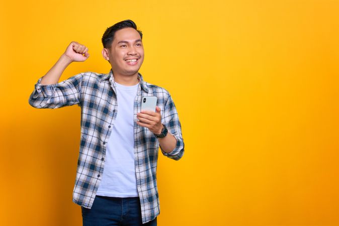 Smiling Asian male holding his phone with his fist raised in celebration with copy space