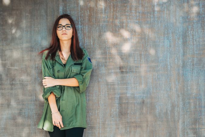 Stylish confident bright woman in glasses leaning on wall and looking at camera