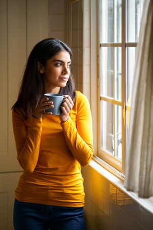 Vertical composition of female gazing out the window with warm beverage