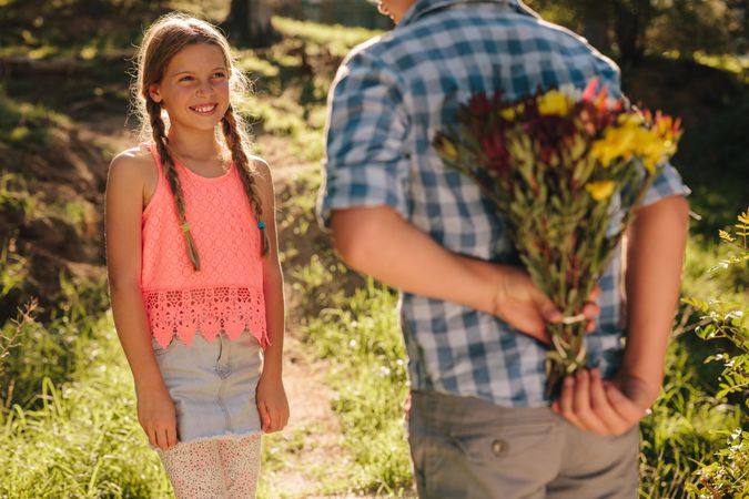 Boy standing in front of his girlfriend in a park holding a bunch of flowers behind him