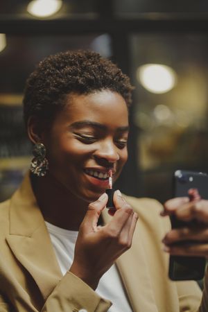 Stylish Black woman rouging lips and holding smartphone on street