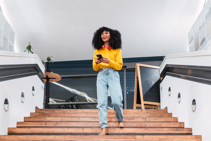 Cheerful stylish Black female walking downstairs and using smartphone in contemporary building