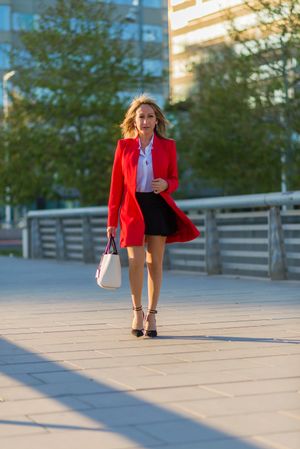 Front view of an elegant woman wearing red jacket, skirt and holding a handbag while walking in the street in a sunny day