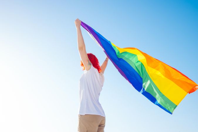 Back view of queer woman waving rainbow flag under blue sky