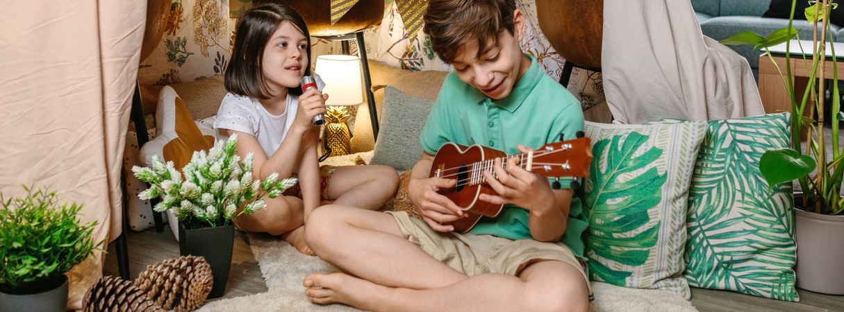 Banner of happy children playing ukulele and singing on handmade shelter tent in living room at home
