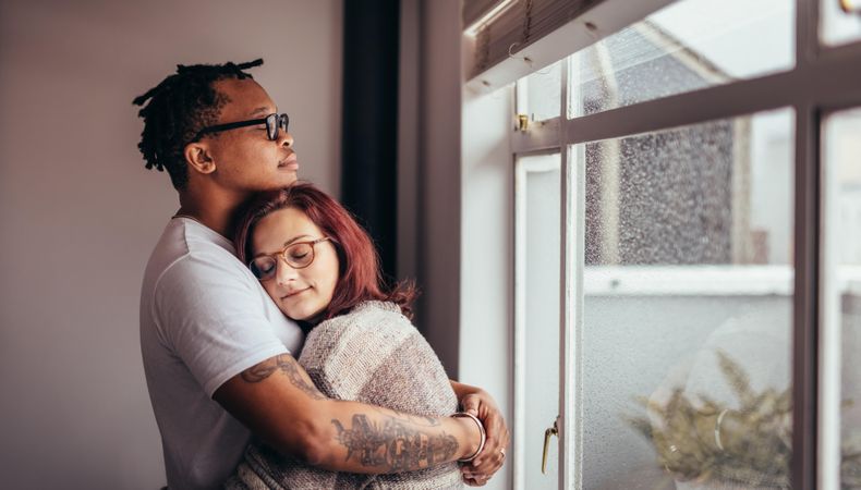 Interracial couple hugging each other while standing near window