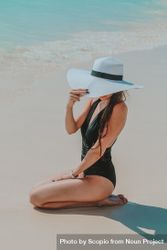 Woman in dark swimsuit wearing light sunhat hat sitting on beach 48EDkb