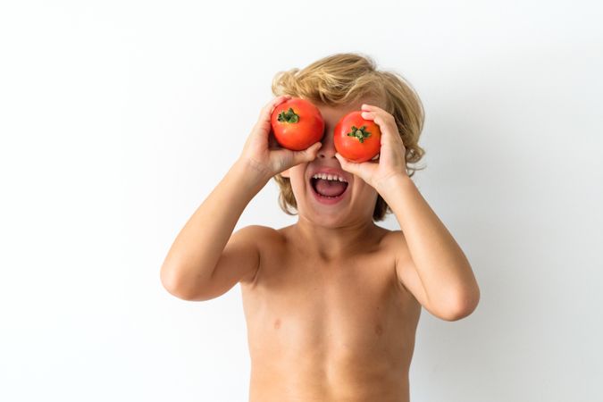 Cheerful boy covering eyes with tomatoes