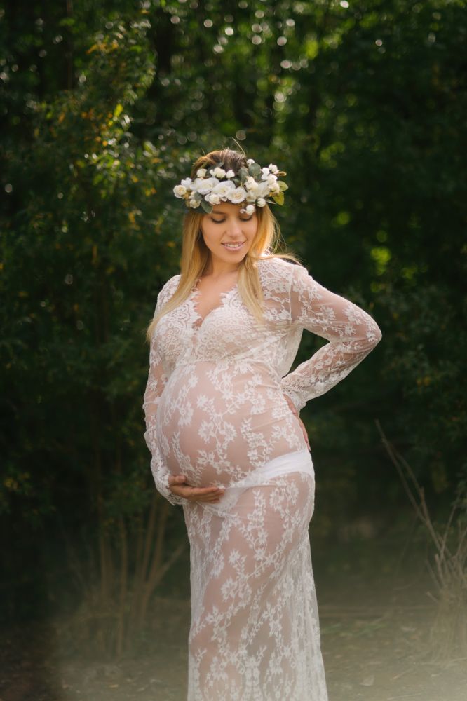 Pregnant woman with closed eyes in white lace dress touching chin while standing in forest