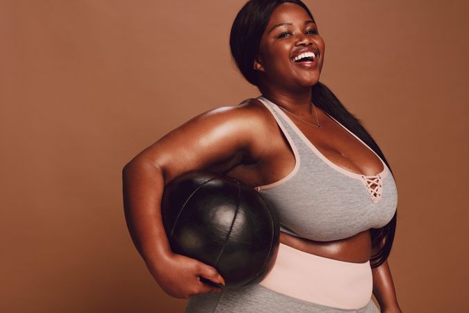 Cheerful plus size Black woman with medicine ball on brown background