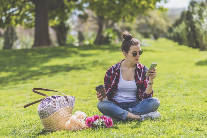 Front view of a young hipster woman wearing sunglasses, sitting on grass in a park while using a mobile phone