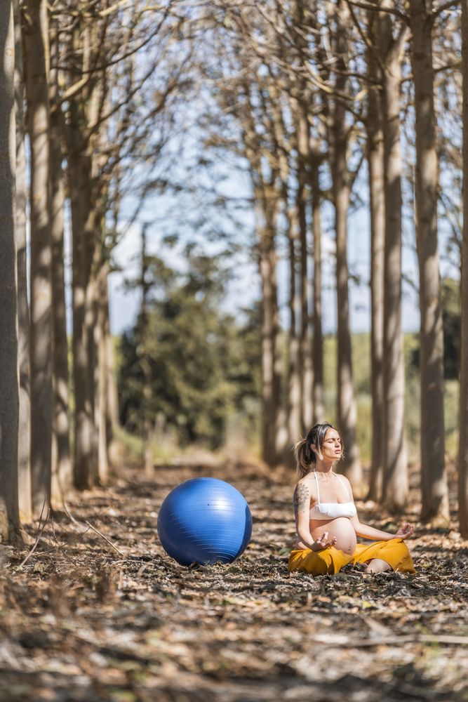 Serene adult pregnant woman meditating while sitting on ground among trees in park during sunny day