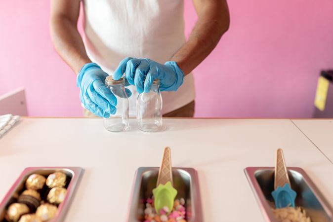 An ice-cream parlor worker is preparing two empty jars to prepare rolls on a counter next to topping trays