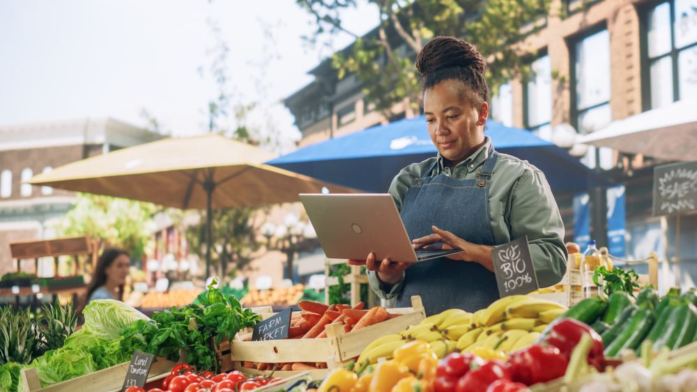 Successful Female Farmer Working on a Laptop Computer, Catching Up with Supplier Online to Plan Future Orders for Her Organic Street Vendor Stall with Sustainable Fruits and Vegetables