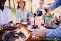 Man pours a glass of red wine at an outdoor table setting among friends O48aXb