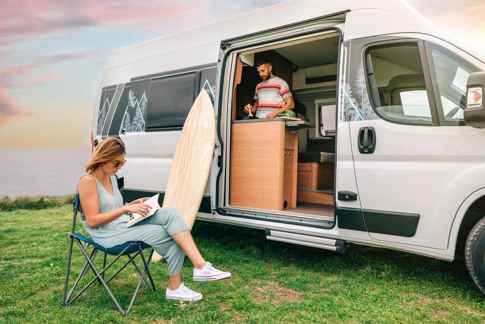 Male cooking inside parked motorhome with female friend sitting outside