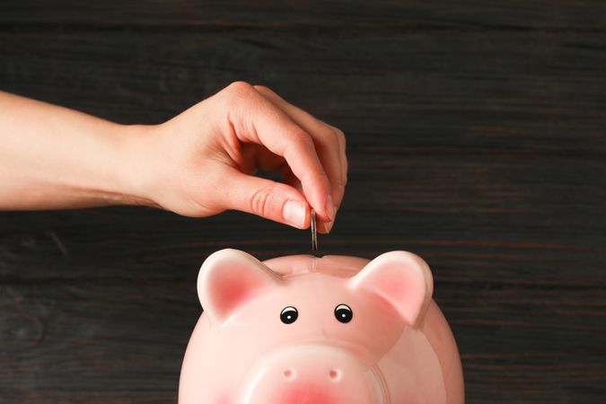 Woman's hand puts a coin in the happy piggy bank on wood table against wooden background, space for text. Finance, saving money