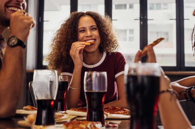 Smiling young woman with curly hair eating pizza with friends at cafe