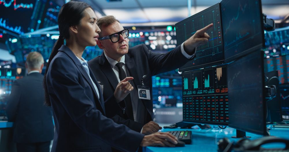 Team of Two Multiethnic Stock Exchange Brokers Using Computer, Chatting About the Asset Management Strategy for an International Business Client, Working in an Office Building with Computer Screens
