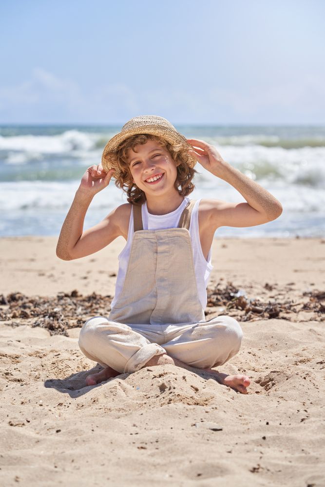 Smiling boy sitting on sandy beach and touching straw hat