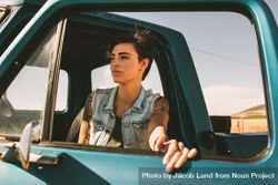 Woman with short hair and tattoos standing near open truck door looking out 4dYeQb