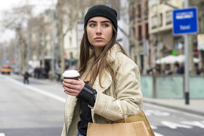 Portrait of female in winter hat crossing the street, with takeaway coffee and shopping bag
