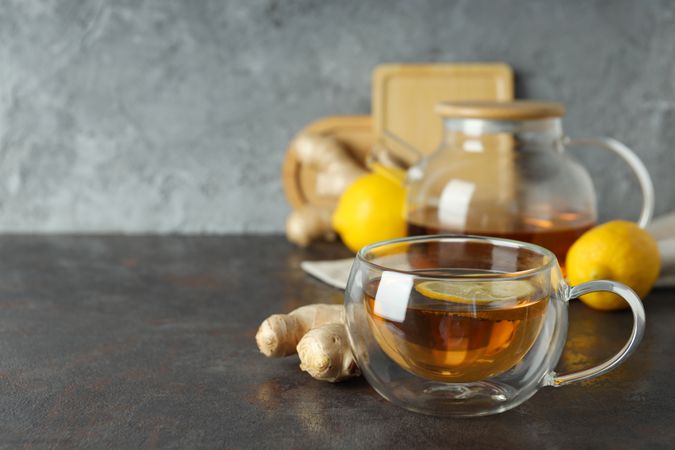 Cup and teapot with tea, lemon and ginger on gray background, space for text