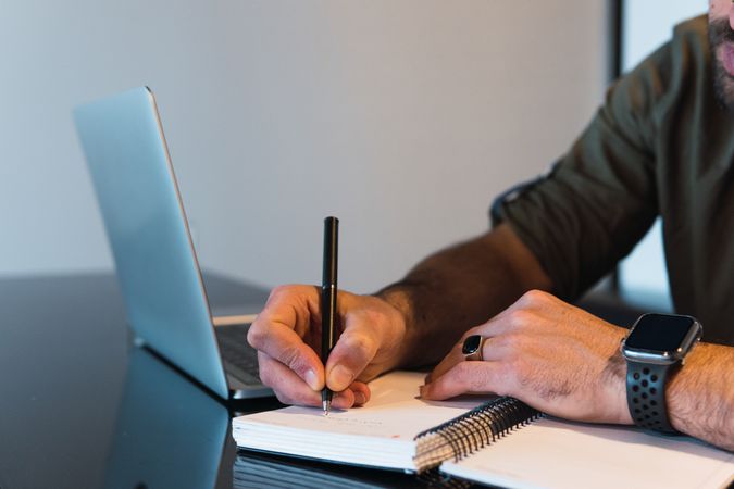 Anonymous man writing in notebook near laptop