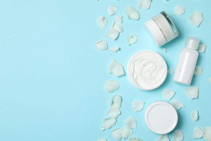 Jars with cream and rose petals on blue background, space for text