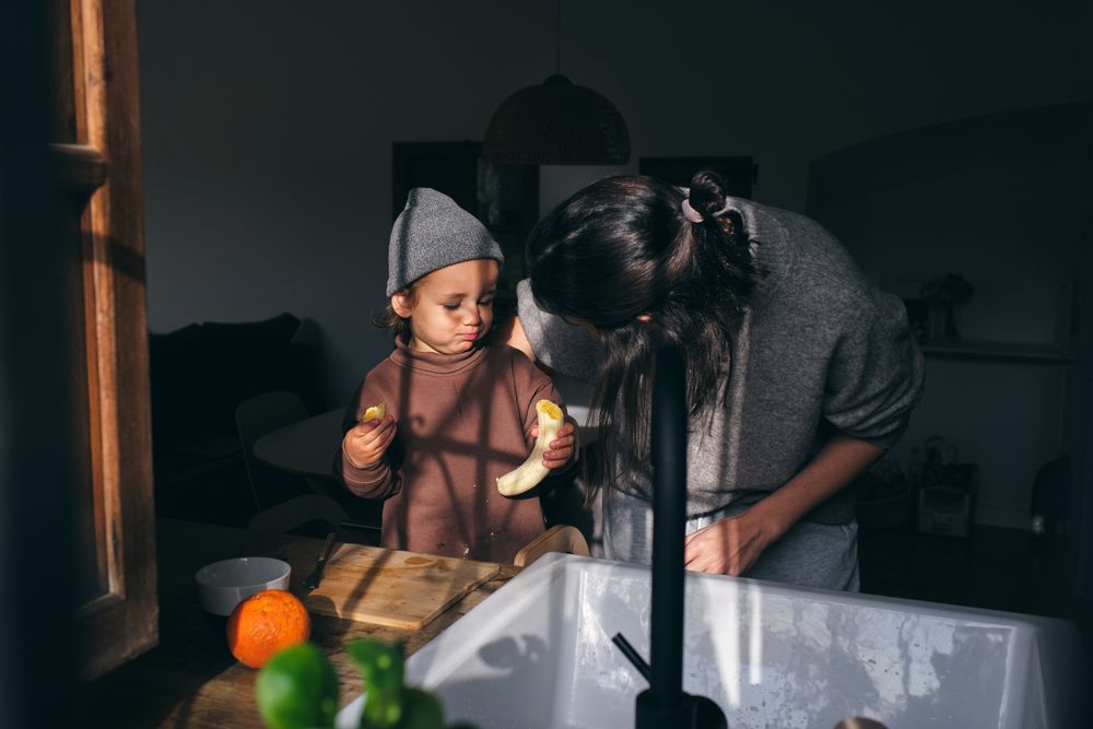 Young mother embracing little son eating banana in kitchen