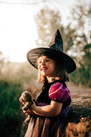 Girl in witch hat with teddy bear in the forest, vertical