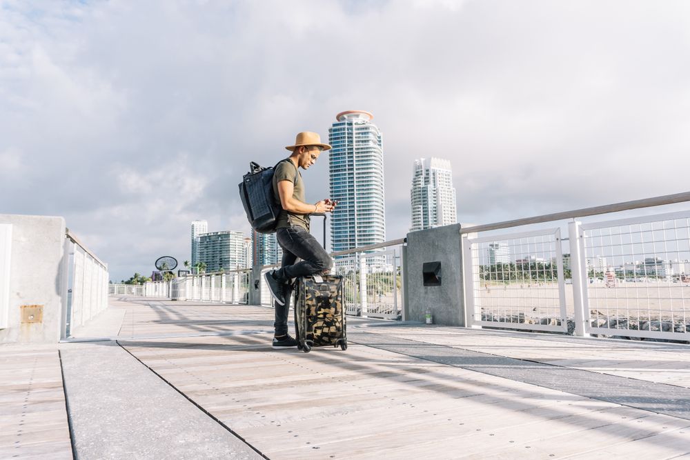 Young man wearing a hat, With A Suitcase and a backpack is searching for directions ion the phone.