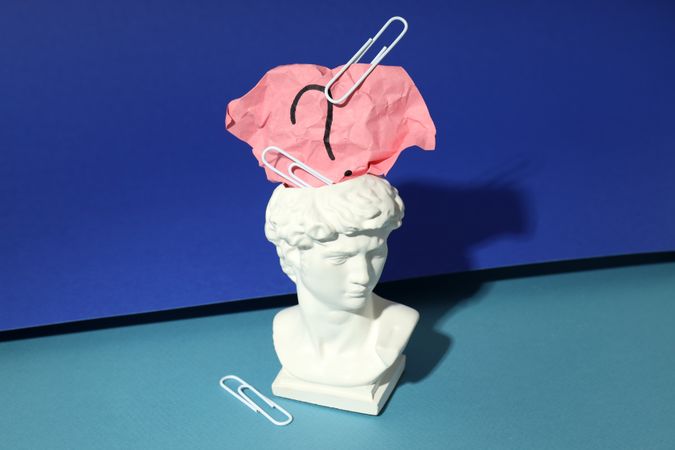 Marble bust of David with crumpled pink post it note with question mark and paper clips, on blue