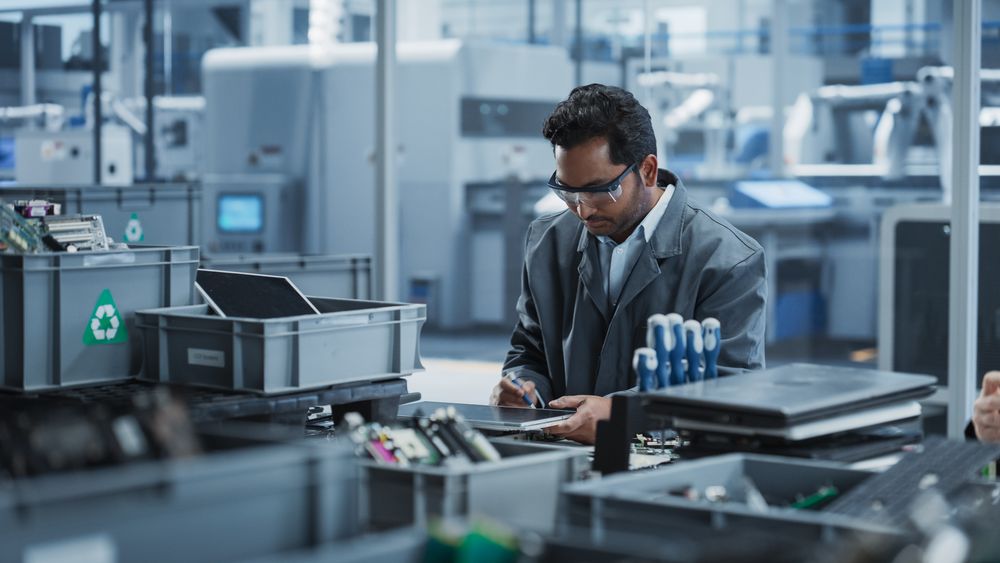Indian Male Worker Disassembling Laptops To Recycle Electronic Components For Microchip Production At An Automated Electronics Factory With Robotic Arms.