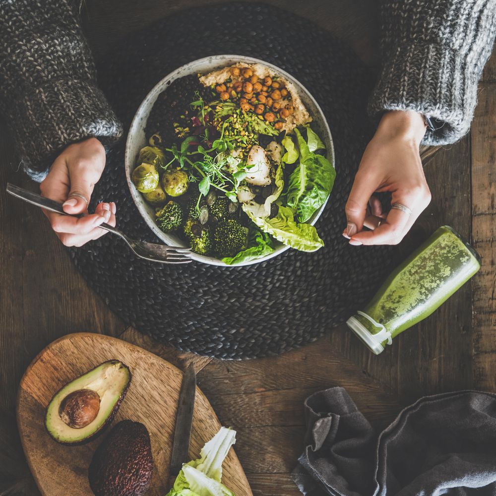 Healthy vegetarian bowl pictured on table with smoothie on side and hands with fork, square crop