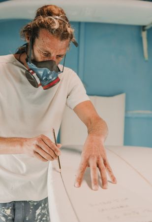 Man in respirator measuring surf board in workshop