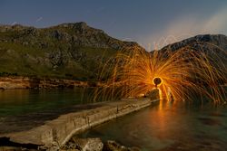 Person with burning circle on stones between water near hills at night