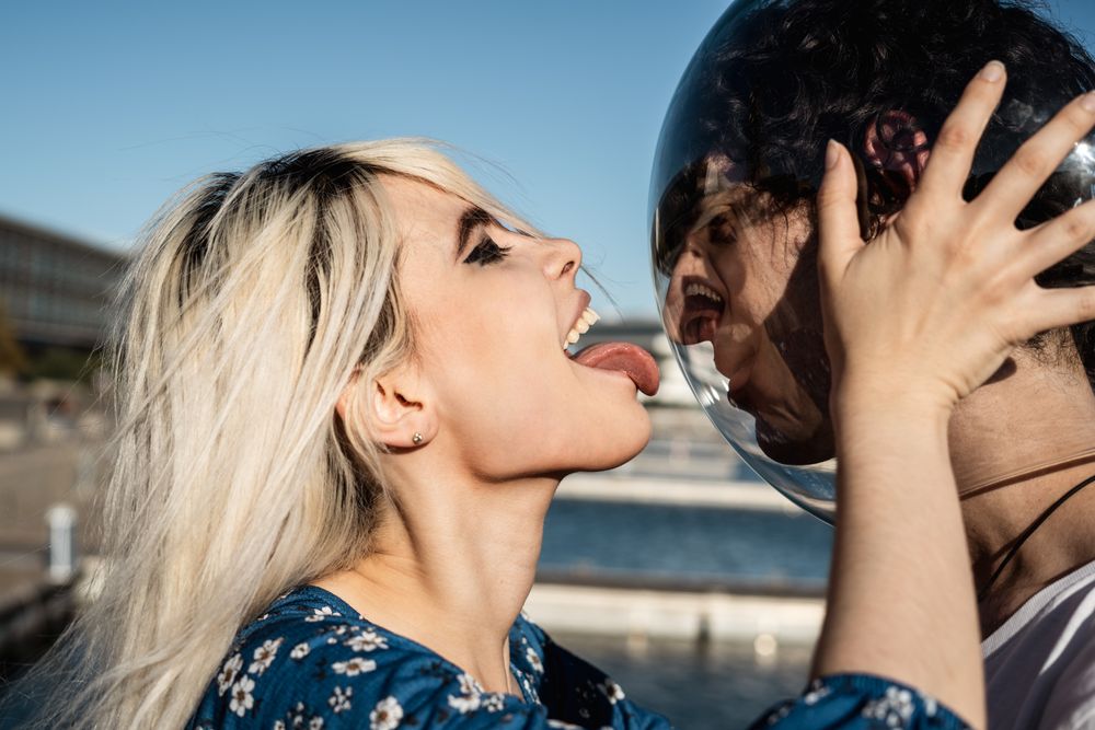 Adult woman touching fish bowl on head of tranquil boyfriend and showing tongue