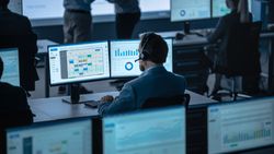 Analyst Reviews Scheduling and Performance Data on Dual Monitors in Busy Office.
