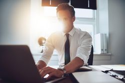 Man in shirt and tie sitting at desk typing at laptop in a sunny office