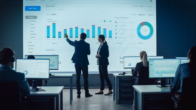 Professionals Analyze Data on a Large Screen in a Modern Office, Focusing on Charts and Graphs.