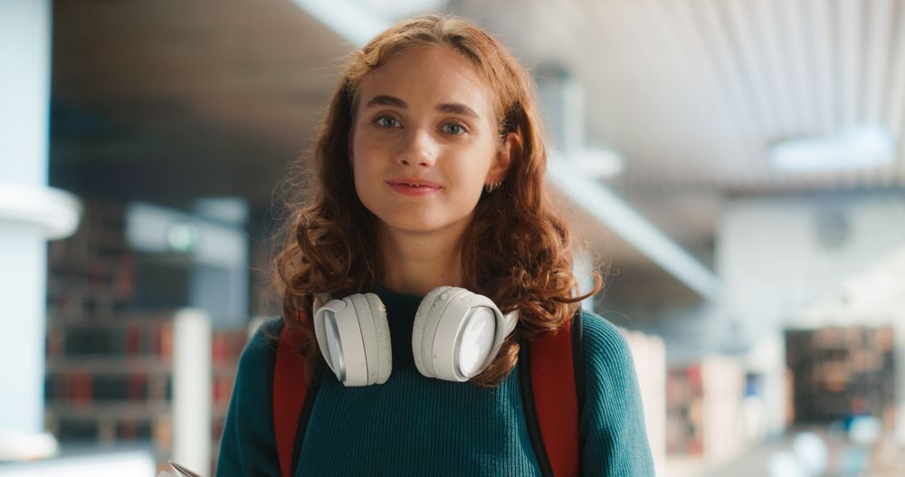 Young Female Student With Headphones Around Neck, Smiling In Modern School Corridor.