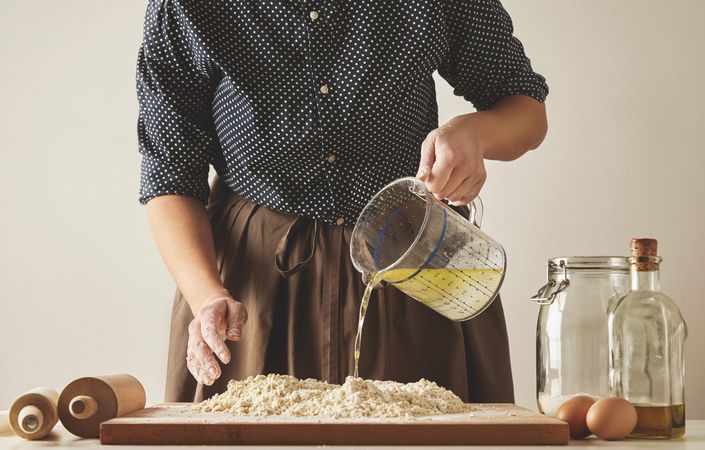 Woman pouring water and oil from measuring cup into pile of dough on bread board