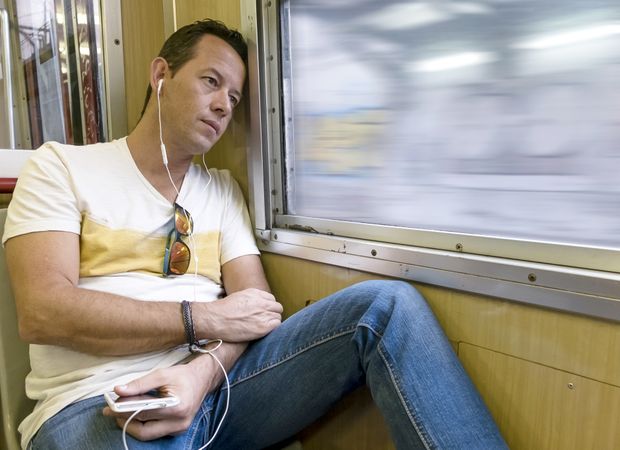 Thoughtful man looking through window inside of suburban train while listening music on earphones
