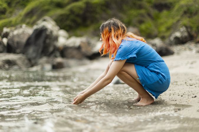 A young female with orange hair, crouching, touches the water with her hands, while gazing at the seashore