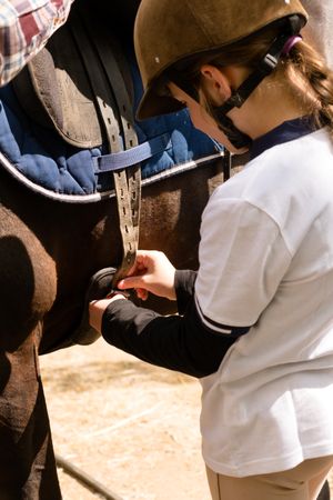 Young rider securing saddle straps on horse.