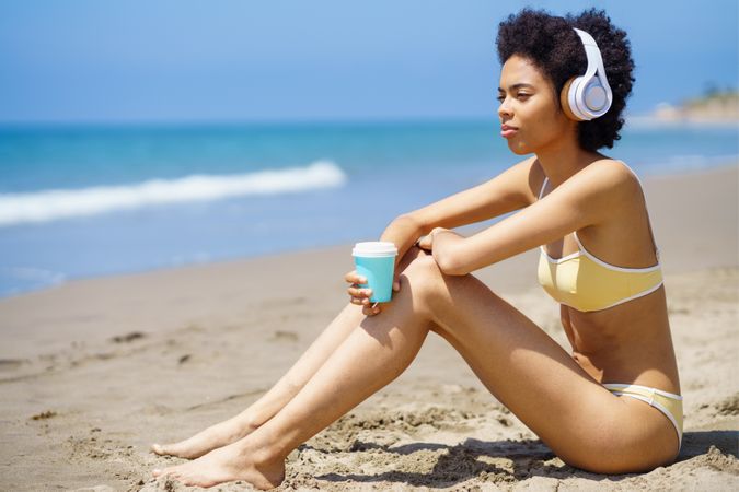 Woman in yellow bikini and headphones sitting on beach with coffee cup