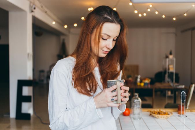 Woman looks down at mug of a warm drink in her apartment